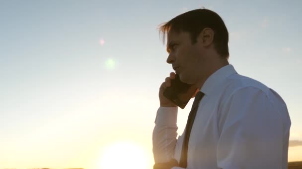 Hombre de negocios hablando en un teléfono inteligente contra el cielo. hombre en un empate con una tableta en el parque al atardecer. hablando de mensaje móvil en el teléfono inteligente. hombre de negocios en una carretera rural con un teléfono móvil — Vídeos de Stock