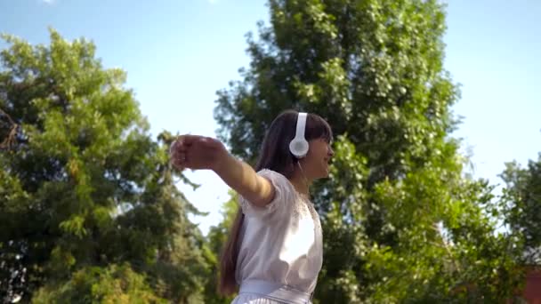 Happy girl in headphones is smiling, holding a smartphone in her hands is dancing and spinning in summer park. beautiful girl listening to music on phone in spring on the street. Slow motion. — Stock Video