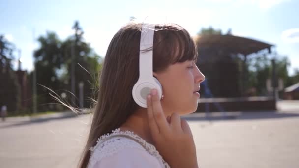 En flicka går längs en stads gata med hörlurar och lyssnar på musik och leenden. flicka i en vit klänning med långt hår färdas runt i staden. Slow motion. tonårs flicka utflykt i staden. — Stockvideo