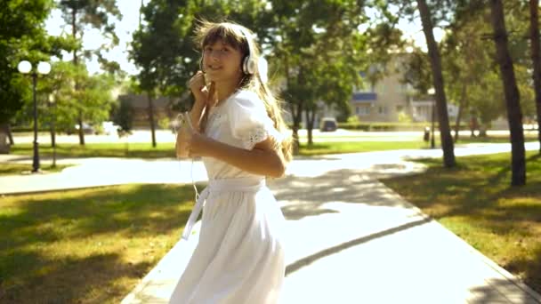 Danças de menina alegres vai no parque no verão na primavera. linda menina dançando, acenando com a mão e chamando para ir com você para viajar. Menina com um telefone em fones de ouvido, ouvir música, sorrindo . — Vídeo de Stock