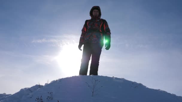 Viajero con mochila desciende de las montañas contra el telón de fondo de la puesta del sol. Turista se encuentra en la cima de la colina nevada en los rayos del sol brillante. concepto de superarse a sí mismo — Vídeos de Stock