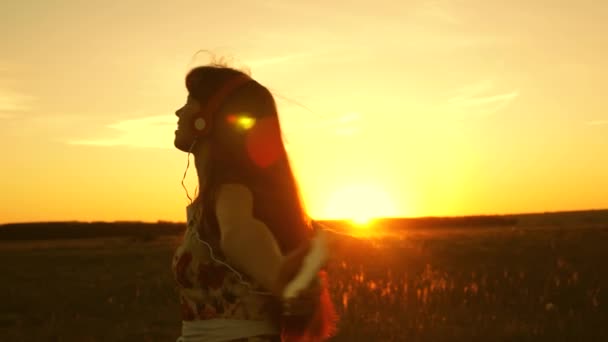 Menina bonita em fones de ouvido e com um giro smartphone em voo sob os raios de um pôr do sol quente. Menina feliz ouvindo música e dançando nos raios de um belo pôr do sol no parque . — Vídeo de Stock