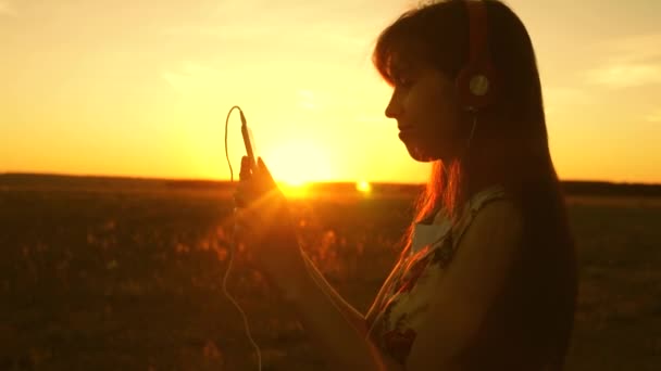 Chica feliz escuchando música y bailando en los rayos de una hermosa puesta de sol. muchacha joven en los auriculares y con un teléfono inteligente toca el dedo al sensor de la tableta selecciona canciones en línea. Movimiento lento . — Vídeos de Stock