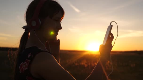 Boldog lány zenét hallgat a smartphone a sugarak egy gyönyörű naplemente. Fiatal lány fejhallgató és a tabletta kiválasztja dalok online. Lassított mozgás. — Stock videók