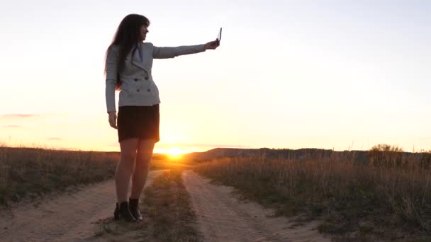 Hermosa mujer de negocios viaja y toma una foto selfie utilizando un teléfono inteligente móvil contra el telón de fondo de la puesta de sol. Chica turística en el campo hace selfie con la tableta. concepto de turismo — Vídeo de stock