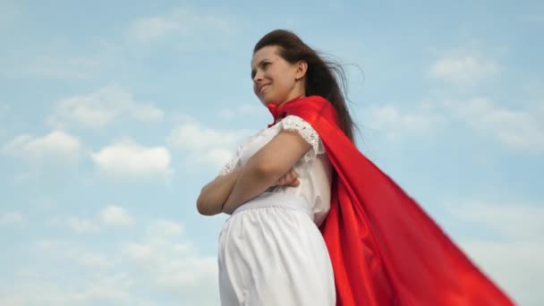 Superhero girl standing on the field in a red cloak, cloak fluttering in the wind. close-up. girl dreams of becoming a superhero. young girl in a red cape dream expression — Stock Video