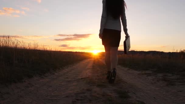 Uma mulher de negócios caminha ao longo de uma estrada rural com um tablet na mão, o sol miertsaet entre as pernas das mulheres. sexy mulher de negócios menina correndo no campo. sexy menina viaja no campo — Vídeo de Stock