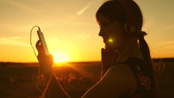 Menina feliz ouvir música no smartphone em raios de um belo pôr do sol de primavera. Movimento lento. jovem com fones de ouvido e com tablet seleciona músicas on-line, no parque no verão em raios de sol . — Vídeo de Stock