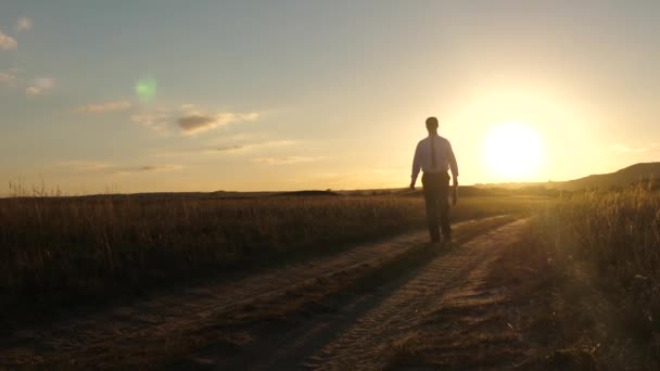 Hombre de negocios con gafas de sol va por el camino del campo con un maletín en la mano. concepto de negocio agrícola. El empresario trabaja en una zona rural. agricultor inspecciona la tierra al atardecer. Movimiento lento . — Vídeo de stock