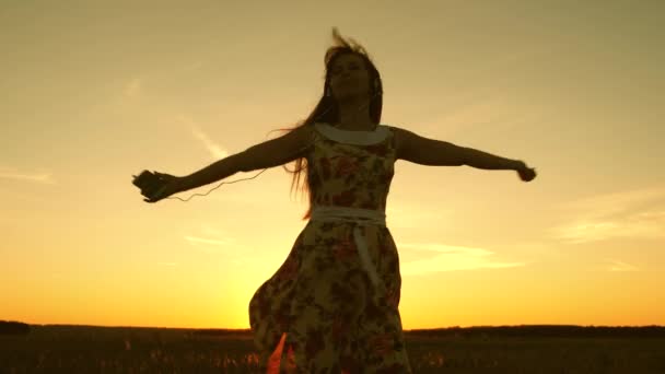 Jeune fille en casque et un smartphone filant en danse sous les rayons d'un coucher de soleil chaud. fille heureuse écoutant de la musique et dansant dans les rayons d'un beau coucher de soleil contre le ciel. Mouvement lent . — Video