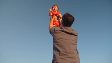 Baba kızını mavi gökyüzüne fırlatıyor. Babam kollarında gökyüzüne karşı küçük bir çocukla oynuyor. mutlu aile gökyüzüne karşı akşam oynarken. baba mutlu bebek, bebek gülümsüyor kusuyor