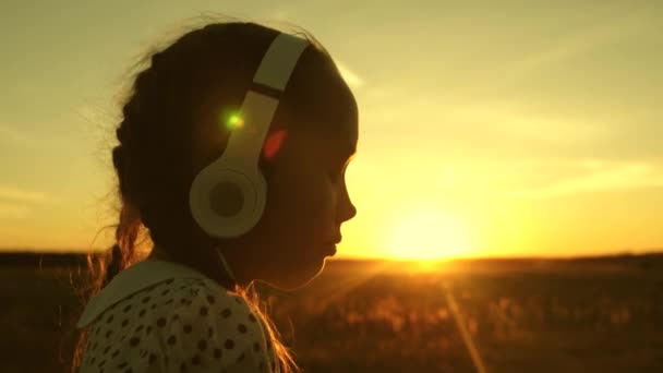 Šťastná dívka poslouchá hudbu na telefonu v paprscích krásného letního slunovratu. dítě se sluchátky a tablet vybírá písně online v létě v parku v paprscích slunce. — Stock video
