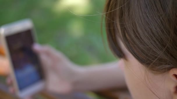 Een jong meisje met behulp van een smartphone is flipping door middel van pagina's in een online winkel in het park op een bankje. Duizendjarige jongedame in het Arboretum, gebaren op het display van de telefoon. — Stockvideo