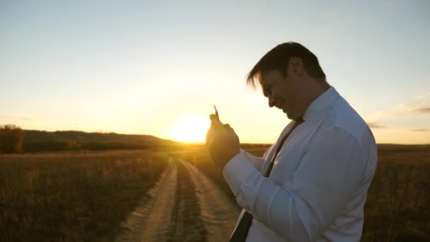 Hombre de negocios jugando juegos de tableta en el parque en los cálidos rayos de la puesta de sol. hombre jugando juegos en el teléfono inteligente. hombre feliz en camisa blanca y corbata juega en línea en la tableta . — Vídeos de Stock