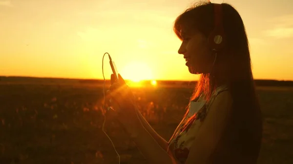 Счастливая девушка слушает музыку и танцует в лучах прекрасного заката. молодая девушка в наушниках и со смартфоном прикасается пальцем к датчику планшета, выбирает песни онлайн. Медленное движение . — стоковое фото