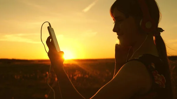 Šťastná dívka naslouchá hudbě na smartphone v paprscích krásného jarního západu slunce. Zpomaleně. mladá dívka se sluchátky a s tabletem vybírá hudbu on-line, v parku v létě v paprscích slunce. — Stock fotografie