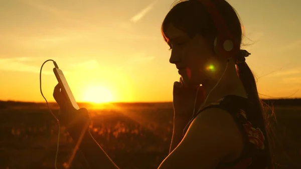 Glückliches Mädchen, das im Strahlen eines schönen Frühlingssonnenuntergangs Musik auf dem Smartphone hört. Zeitlupe. junges Mädchen mit Kopfhörern und Tablet wählt Lieder online aus, im Sommer im Park in Sonnenstrahlen. — Stockfoto