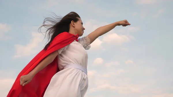 Bela menina super-herói de pé em um campo em um manto vermelho, vacilante manto no vento. menina sonha em se tornar um super-herói. Movimento lento. jovem joga em um manto vermelho com expressão de sonhos . — Fotografia de Stock