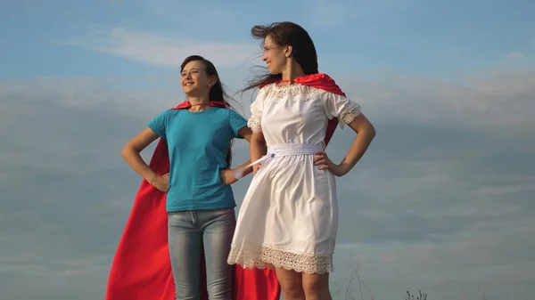 Dvě dívky v červených pláštích superhrdinů se postaví proti modrému nebi, vítr nafouká plášť. Máma s dcerou hrají superhrdiny. šťastný rodinný koncept — Stock fotografie