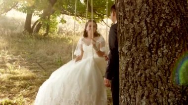 Mutlu damat yazın parkta gelini salıncakta sallıyor. erkek ve kız yaz ormanında bir meşe dalı üzerinde salıncak. mutlu aile konsepti. aşık bir çiftin takım çalışması.