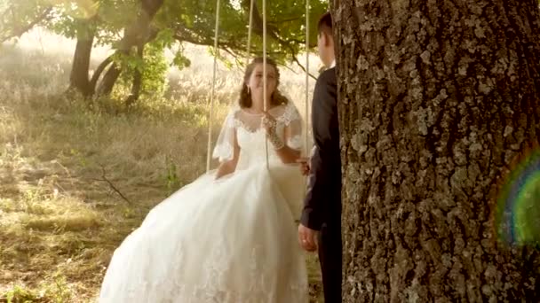 Lo sposo felice che oscilla su un'oscillazione la sposa nel parco in estate. ragazzo e ragazza oscillano su un ramo di una quercia nella foresta estiva. felice concetto di famiglia. lavoro di squadra di una coppia innamorata . — Video Stock