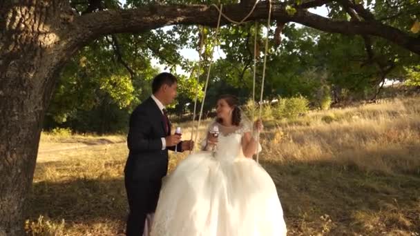 Lo sposo felice che oscilla su un'oscillazione la sposa nel parco in estate. ragazzo e ragazza oscillano su un ramo di una quercia nella foresta estiva. felice concetto di famiglia. lavoro di squadra di una coppia innamorata . — Video Stock