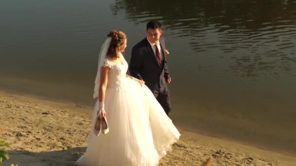 Das glückliche Brautpaar spaziert barfuß im Sand am Flussufer entlang. Verliebtes Paar geht Hand in Hand am Strand. Glückliches Familienkonzept. Frischvermählte Flitterwochen am Strand. Teamwork eines verliebten Paares. — Stockvideo