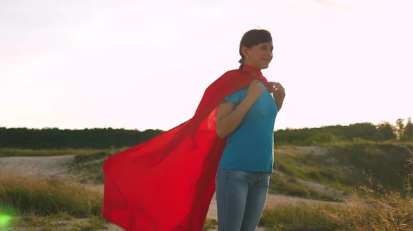 Bela menina super-herói em pé no campo em um manto vermelho, vacilante manto no vento. Movimento lento. Uma jovem sonha em se tornar uma super-heroína. menina caminha em um manto vermelho expressão de sonhos — Fotografia de Stock