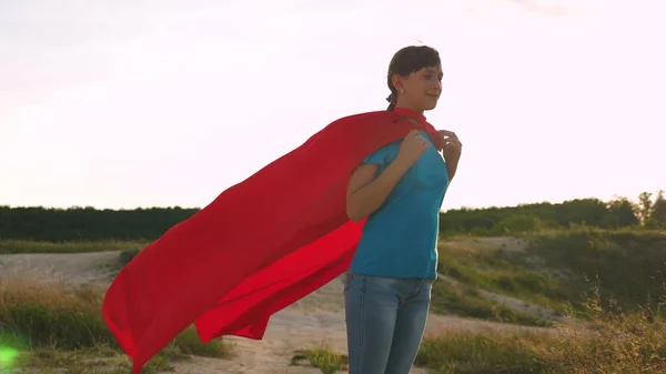 Mooi meisje superhero staande op het veld in een rode mantel, mantel wapperen in de wind. Slow-motion. Een jong meisje droomt ervan om van een superheld. meisje loopt in de expressie van een rode mantel van dromen — Stockfoto