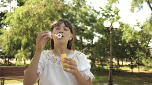 Young girl playing in the park and blowing bubbles into the camera lens. Slow motion. Beautiful girl blowing soap bubbles in the park in spring, summer and smiling. — Stock Video