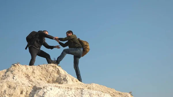 Turistas dão a mão uns aos outros, subindo para o topo da colina. equipe de viajantes do sexo masculino vai para a vitória e sucesso. trabalho de equipa de pessoas de negócios. três alpinistas escalam um após o outro na rocha branca . — Fotografia de Stock