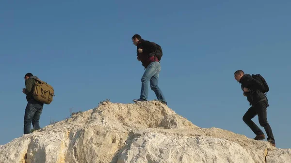 Turistas passam para o topo da colina contra o céu. trabalho de equipa de pessoas de negócios. três alpinistas escalam um após o outro na rocha branca. equipe de viajantes do sexo masculino vai para a vitória e sucesso . — Fotografia de Stock