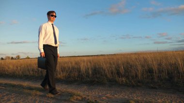 işadamı güneş gözlüğü elinde bir çanta ile ülke yolda gidiyor. Girişimci bir kırsal bölgede çalışıyor. bir çiftçi kara gün batımında olup olmadığını denetler. tarıma dayalı işletme kavramı.