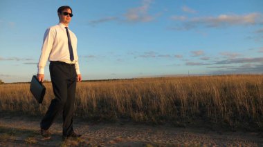işadamı güneş gözlüğü elinde bir çanta ile ülke yolda gidiyor. Girişimci bir kırsal bölgede çalışıyor. bir çiftçi kara gün batımında olup olmadığını denetler. tarıma dayalı işletme kavramı.