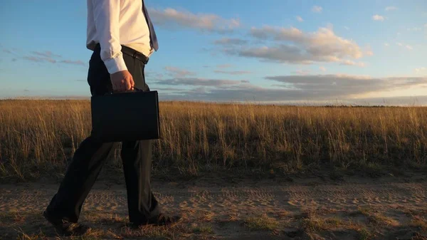 Uomo d'affari in occhiali da sole va lungo la strada di campagna con una valigetta in mano. L'imprenditore lavora in una zona rurale. un agricoltore ispeziona la terra al tramonto. concetto di impresa agricola . — Foto Stock