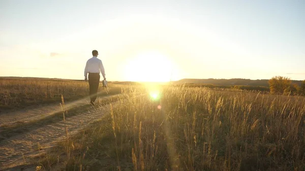 Uomo d'affari in occhiali da sole va lungo la strada di campagna con una valigetta in mano. L'imprenditore lavora in una zona rurale. un agricoltore ispeziona la terra al tramonto. concetto di impresa agricola . — Foto Stock