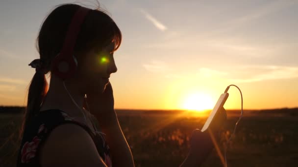 Chica feliz viaja y escucha música en su teléfono inteligente en los rayos de una hermosa puesta de sol. Chica joven en el parque con auriculares y con una tableta selecciona canciones en línea . — Vídeo de stock