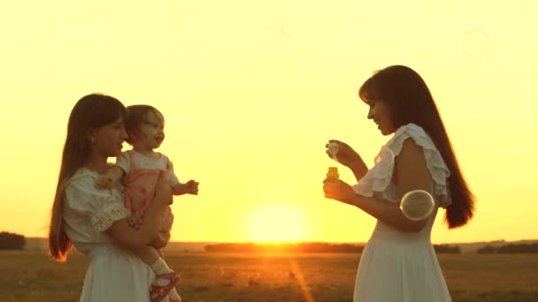 Glückliche Mutter, die mit Kindern spielt und Seifenblasen pustet. Töchter und Mutter pusten bei Sonnenuntergang Blasen in den Park. Zeitlupe. Glückliches Familienkonzept. Baby, Schwester und Mutter spielen in der Sonne. — Stockvideo