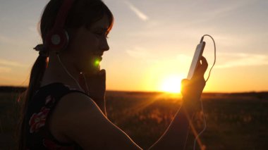 Güzel bir gün batımı ışınları bir akıllı telefonda müzik dinleyen mutlu kız. Kulaklıkta genç kız ve tablet ile online şarkıları seçer. Yavaş hareket.