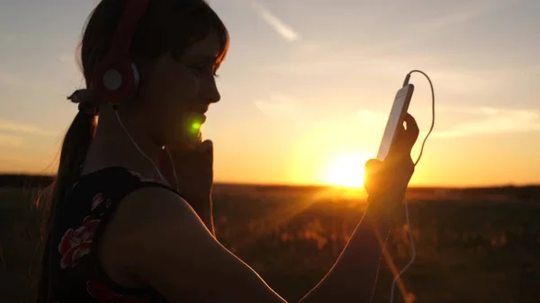 Ragazza felice che ascolta musica su uno smartphone nei raggi di un bellissimo tramonto. Giovane ragazza in cuffia e con il tablet seleziona canzoni online. Rallentatore . — Foto Stock