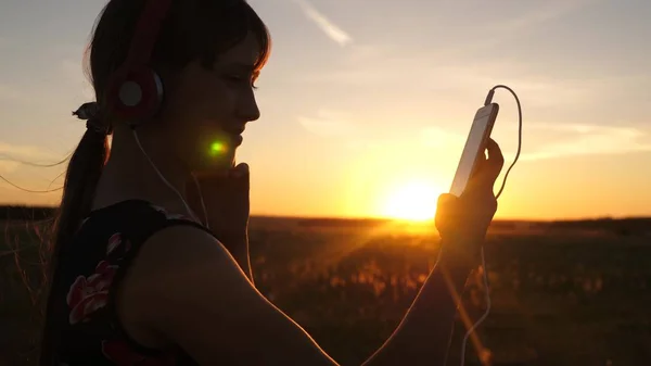 Glückliches Mädchen, das Musik auf einem Smartphone im Strahlen eines schönen Sonnenuntergangs hört. junges Mädchen mit Kopfhörer und Tablet wählt Lieder online aus. Zeitlupe. — Stockfoto