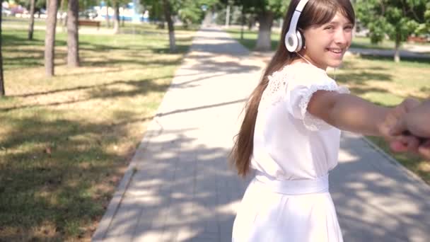 Venha atrás de me.girl em fones de ouvido sorri e leva o homem amado pela mão no parque um dia de primavera. trabalho de equipa de um casal amoroso. menina e menino viajar para a música de mãos dadas. Movimento lento . — Vídeo de Stock