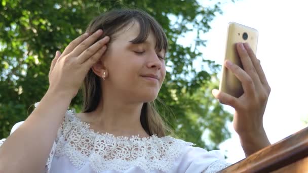 Chica adolescente navega fotos en un teléfono inteligente en un banco. comprobar correo electrónico en línea. hermosa chica escribe en el teléfono inteligente una carta en el parque en primavera, verano. Chica joven charlando en una tableta con amigos . — Vídeo de stock