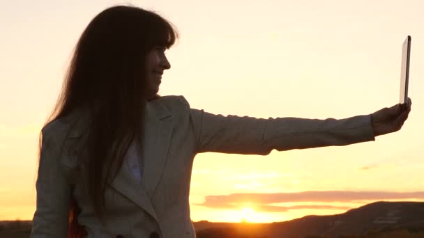 Turista menina no campo faz selfie com tablet. bela mulher de negócios viaja e tira uma foto selfie usando um smartphone móvel contra o pano de fundo do pôr do sol. conceito de turismo — Vídeo de Stock