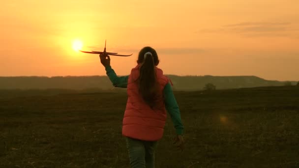 Mutlu kız alan üzerinde bir günbatımı arka planda bir oyuncak uçak ile çalışır. Mutlu bir aile kavramı. Bir çocuk oyuncak uçak çalar. Kız uçmayı ve pilot olmayı hayal ediyor. Yavaş — Stok video