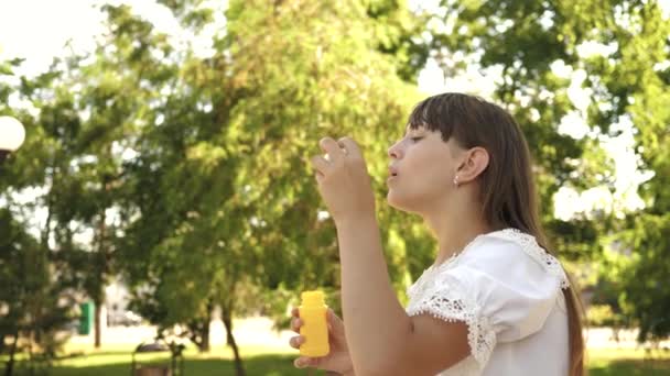 Chica feliz soplando hermosas burbujas de jabón en el parque en primavera, verano y sonriendo. En cámara lenta. niña viajando por la ciudad en el parque . — Vídeo de stock