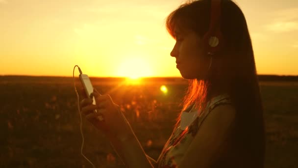 Meisje luisteren naar muziek en dansen in de stralen van een prachtige zonsondergang. een jong meisje in een koptelefoon en met een smartphone raakt de vinger aan de sensor van de Tablet selecteert nummers online. Slow Motion. — Stockvideo