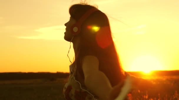 Όμορφο κορίτσι στα ακουστικά και με ένα smartphone στροβιλίζεται στην πτήση κάτω από τις ακτίνες του ένα ζεστό ηλιοβασίλεμα. Ευτυχισμένο κορίτσι ακούγοντας μουσική και χορεύοντας στις ακτίνες ενός όμορφου ηλιοβασιλέματος στο πάρκο. — Αρχείο Βίντεο