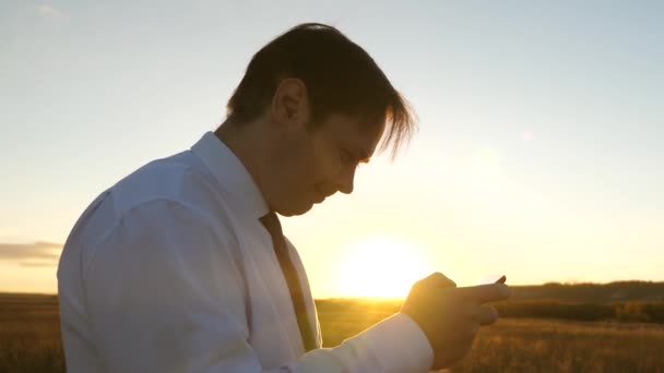 Hombre de negocios jugando juegos de tableta en el parque en los cálidos rayos de la puesta de sol. hombre jugando juegos en el teléfono inteligente. hombre feliz en camisa blanca y corbata juega en línea en la tableta . — Vídeos de Stock