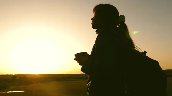 Mujer viajera silueta, se encuentra en la cima de una colina bebiendo café en vidrio de termos. turista chica bebe té caliente y mira al sol. Descanso aventurero después de alcanzar la meta. concepto de libertad . —  Fotos de Stock
